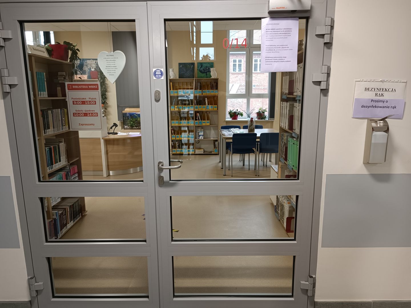 Zdjęcie przedstawia wejście do biblioteki z dozownikiem z płynem do dezynfekcji rąk umieszczonym na śzianie obok drzwi