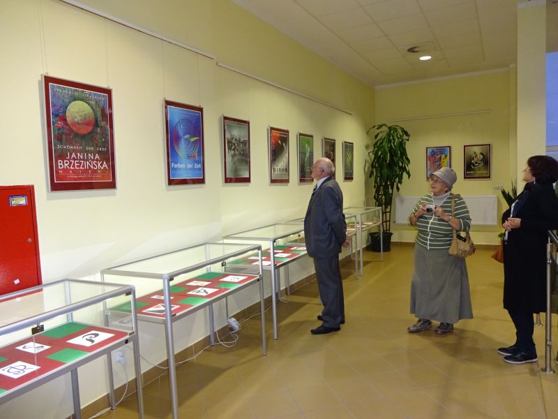Trzy osoby oglądające wystawę, na ścianach wiszą plakaty, w gablotach prezentowane są znaki graficzne