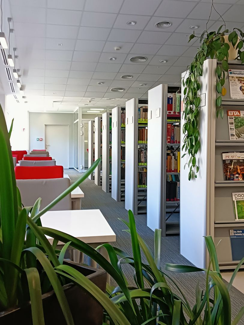 Widok na rząd stolików pod oknem i regały w Bibliotece Wydziału Budownictwa i Inżynierii Środowiska